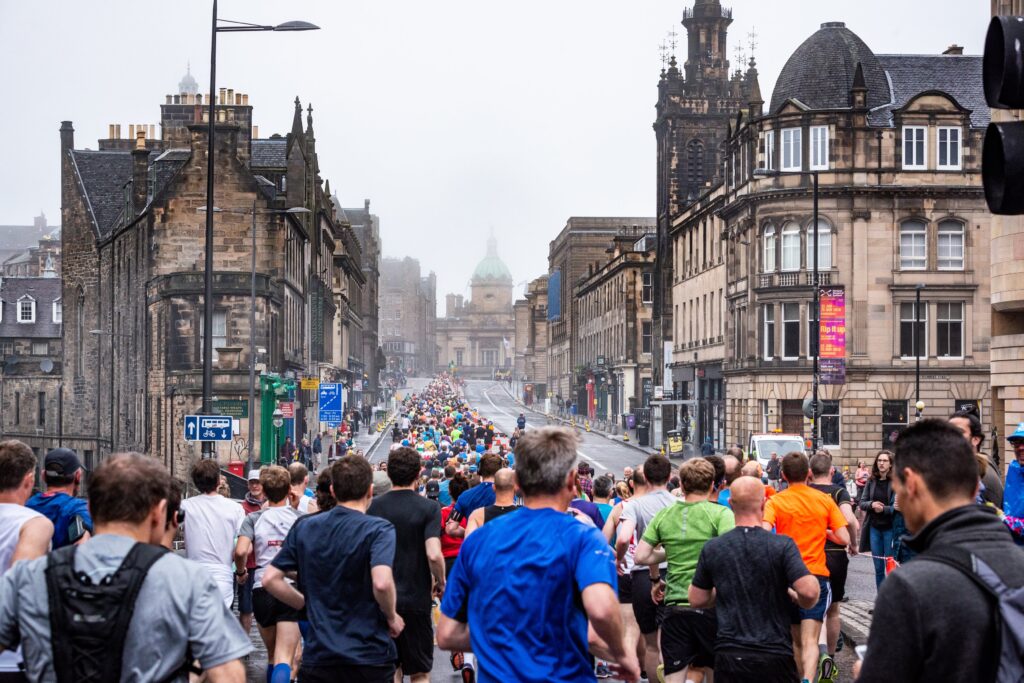 A large group of runners with backs to the camera, running through a street in Edinburgh.