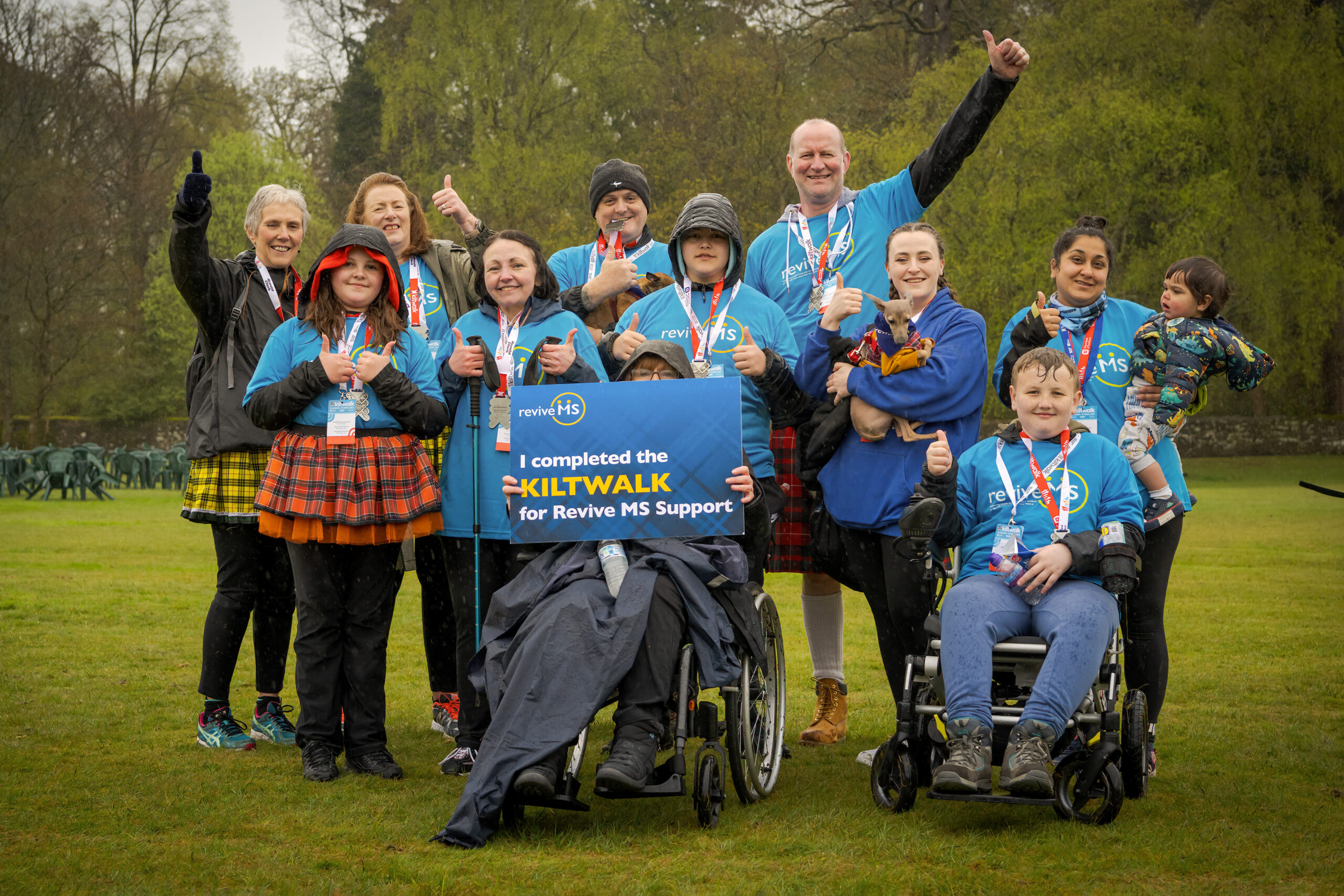 A group of twelve people stand outside in a field with trees in the background. They are all wearing blue Revive t-shirts. Two people at the front are sitting in wheelchairs, one holds a blue sign that reads I completed the Kiltwalk for Revive MS Support. On the right of the image one woman is holding a small dog and the women next to her has a baby in her arms.