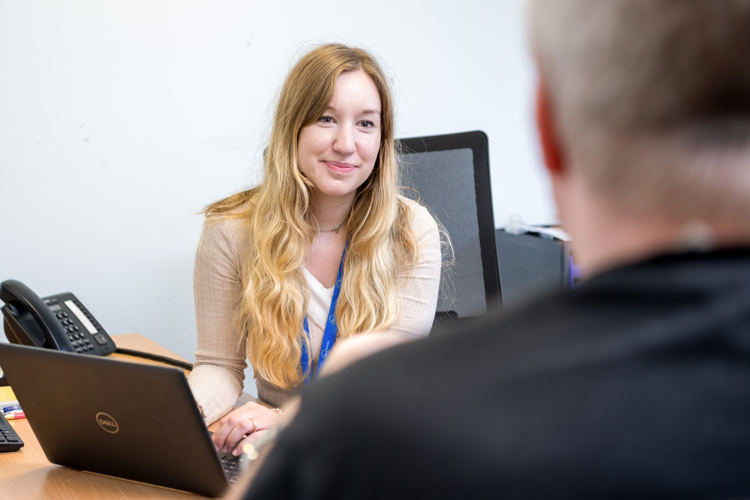 A young woman with long blond hair and a cream top is speaking to a man who has his back to the camera. She is taking notes on a laptop and smiling gently.