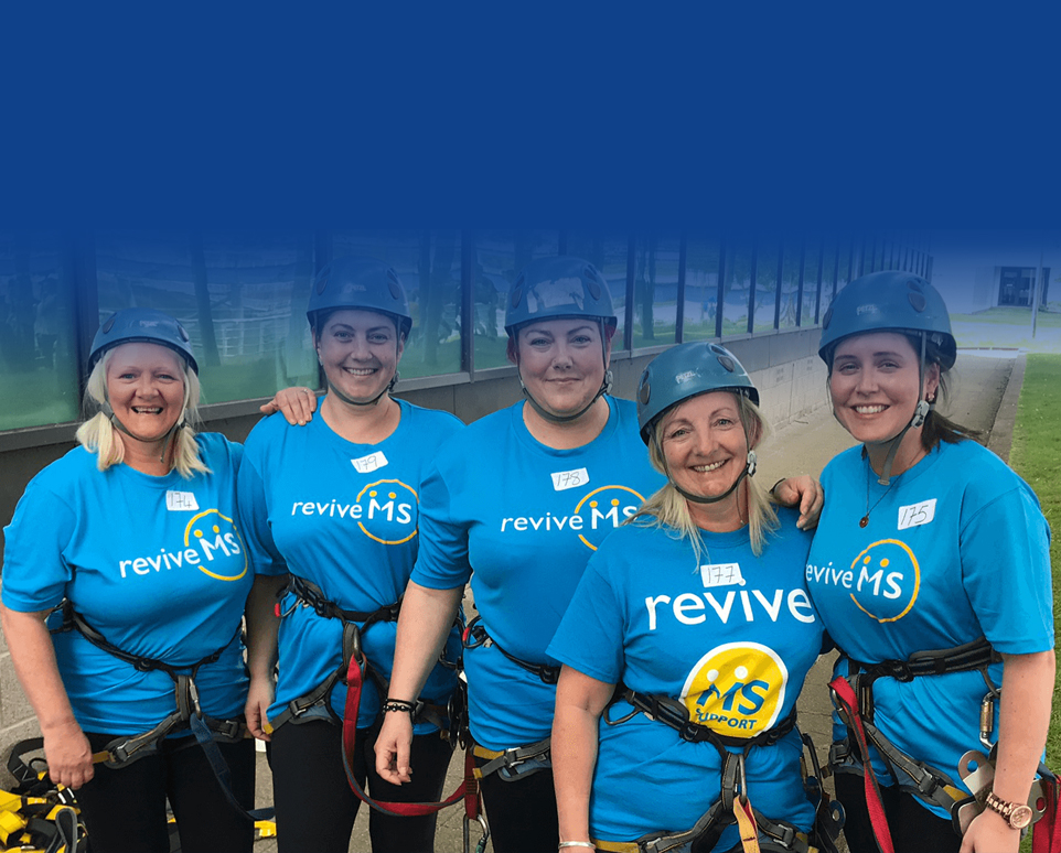 A group of five people in blue Revive t-shirts are wearing hard hats and harnesses. They are smiling and looking directly at the camera.