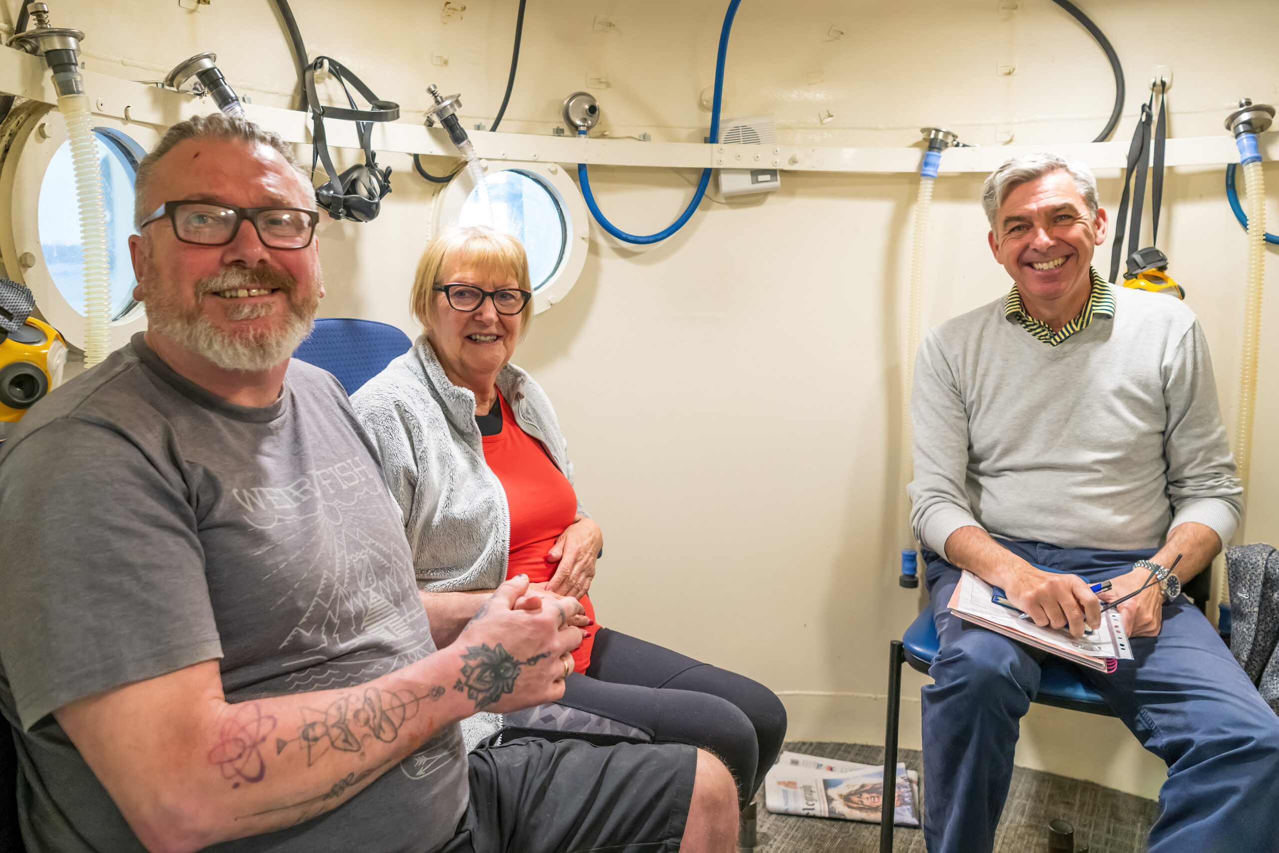 Three people are sitting in the oxygen chamber. They are smiling and looking at the camera.