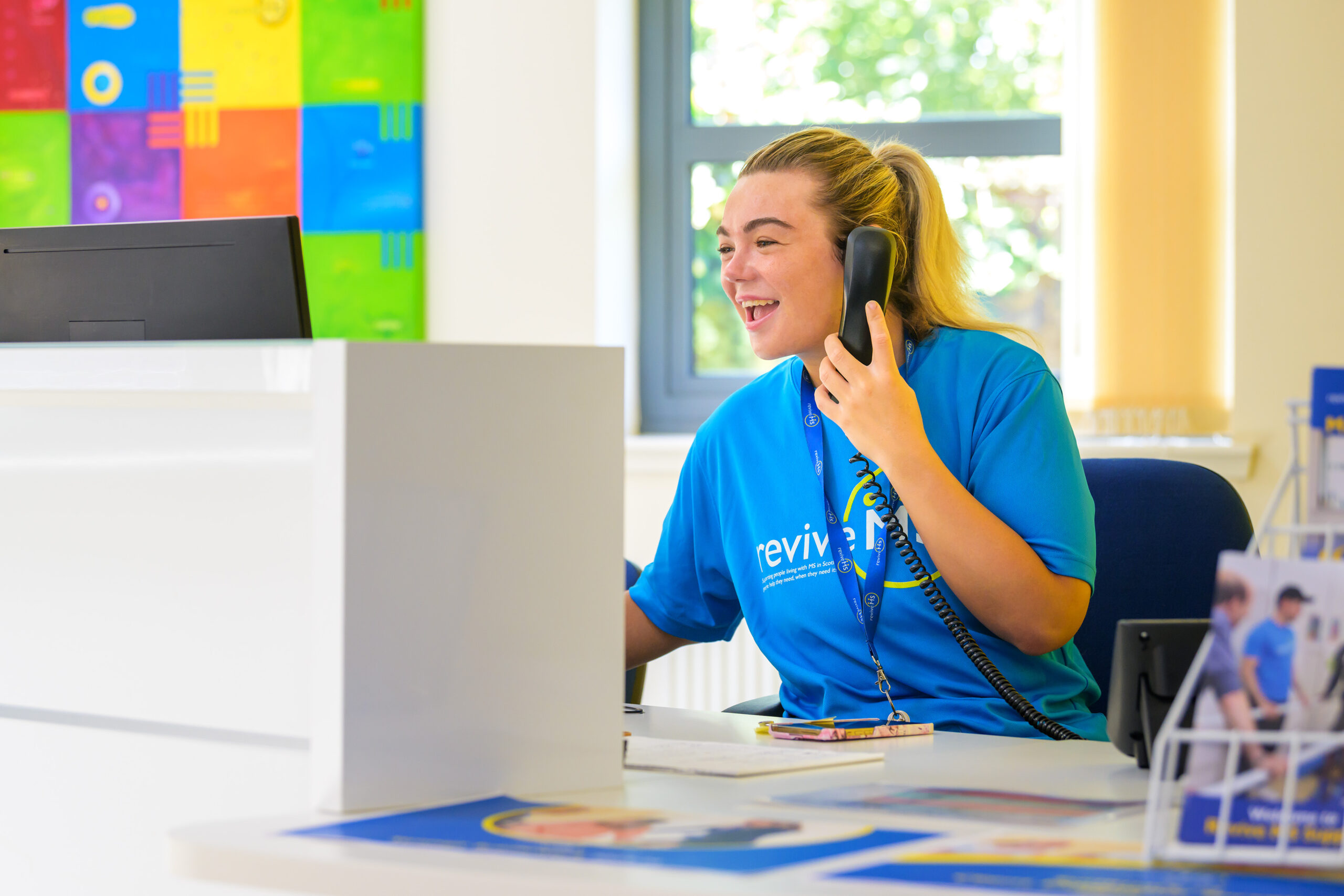 A young woman with long blonde hair is talking on the phone and smiling. She wears a bright blue t-shirt with the Revive logo on it.