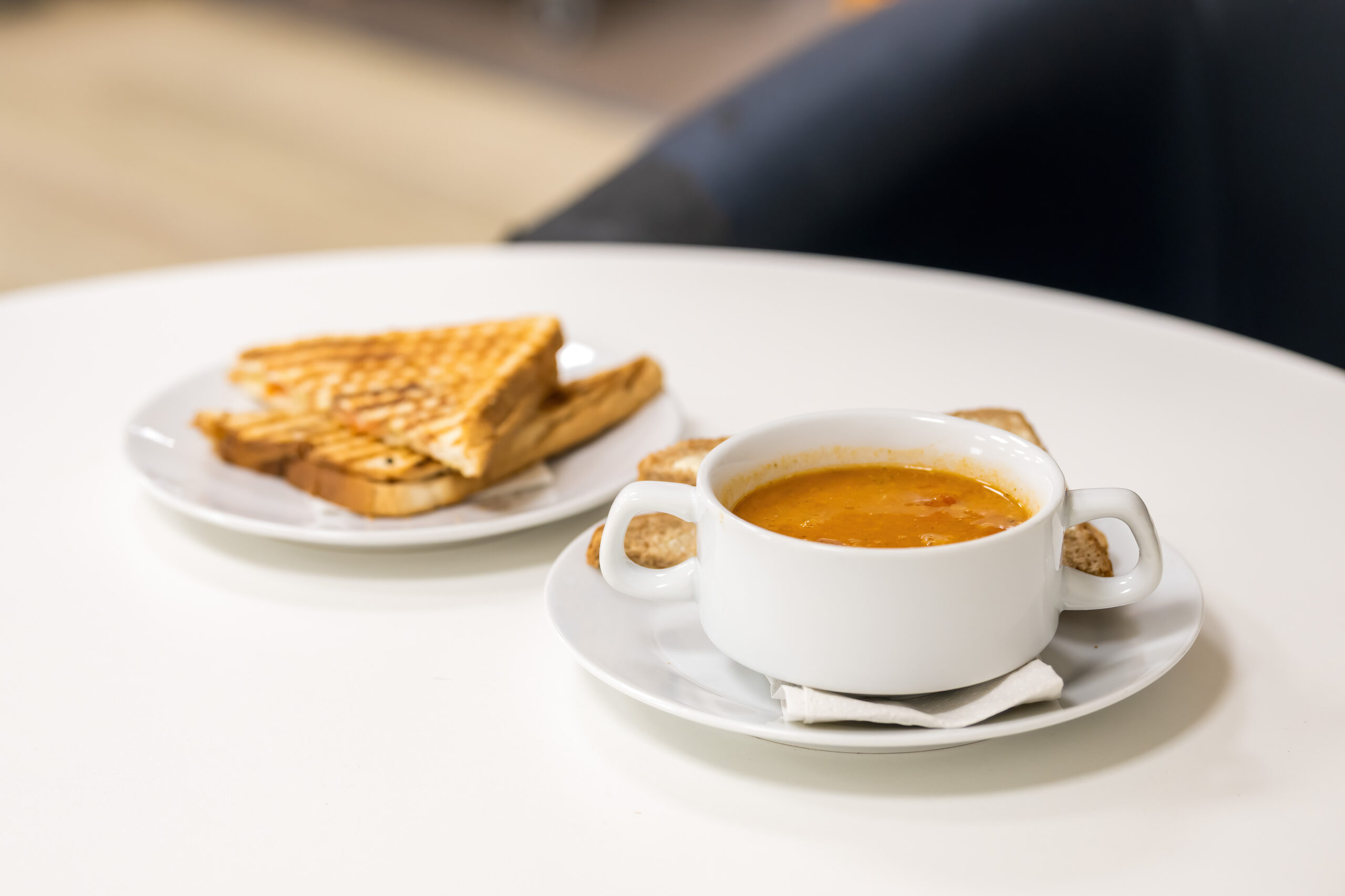 A bowl of soup and a plate with a toastie sits on a white table.