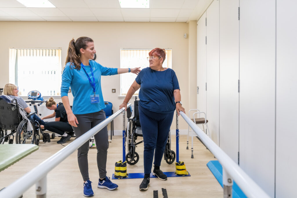 The image shows the Female Revive Physiotherapist putting her arm around a Female client. The client is walking in between parallel walking bars. Both women are smiling at each other.