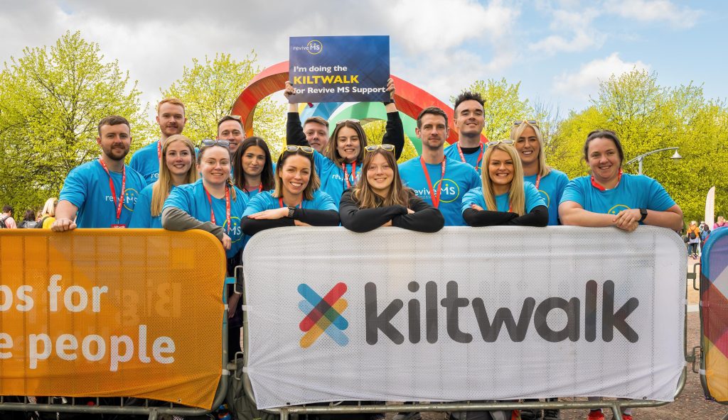 A group of smiling Team Revive MS Support walkers wearing blue Revive MS Support t-shirts stand behind a Kiltwalk barrier at the event start line. They are posing together under a colourful Kiltwalk archway on a bright day with trees in the background.