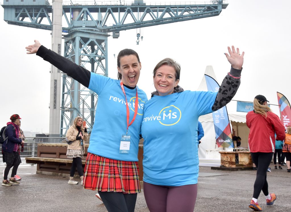 Two smiling women wearing blue Revive MS Support t-shirts pose with their arms raised at the Glasgow Kiltwalk. One wears a red tartan skirt, and the Finnieston Crane can be seen in the background, with other walkers and event tents nearby